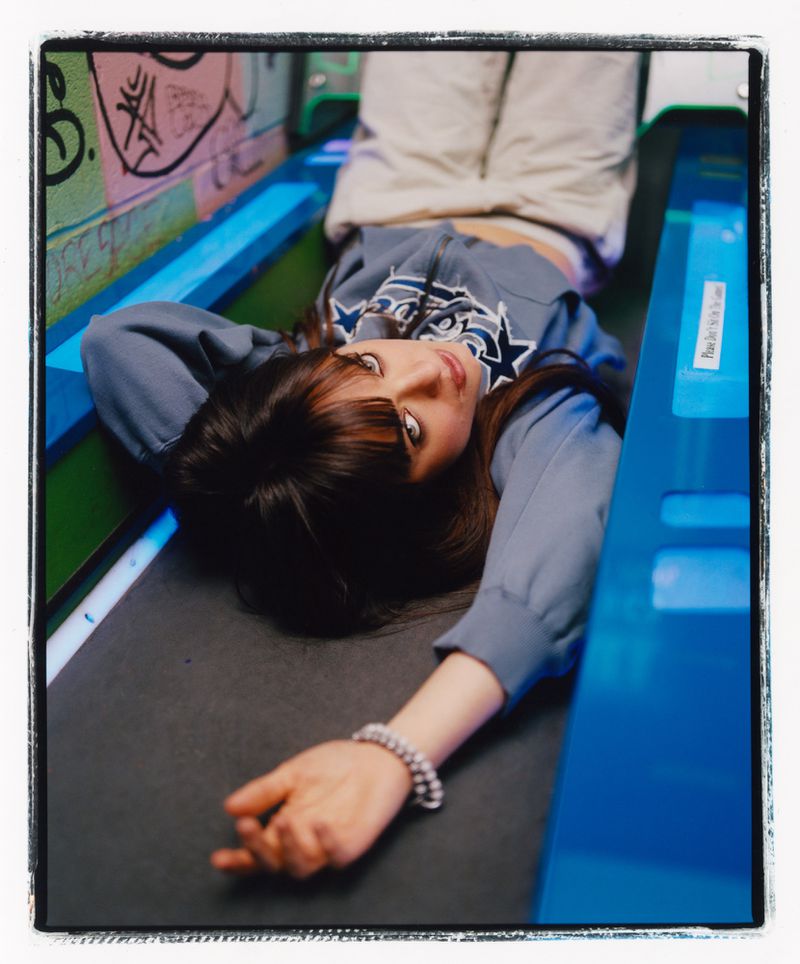 Brunette woman with bangs lies on her back, looking up at the camera wearing a grey sweatshirt and light pants in an indoor setting.