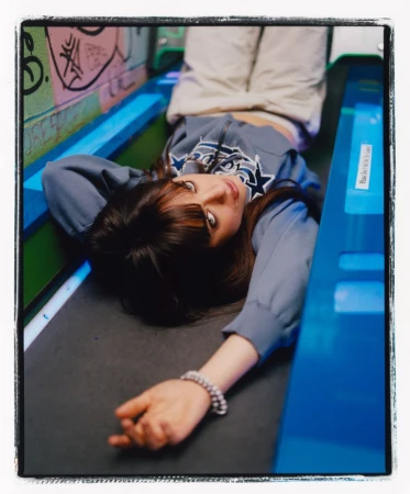 Brunette woman with bangs lies on her back, looking up at the camera wearing a grey sweatshirt and light pants in an indoor setting.