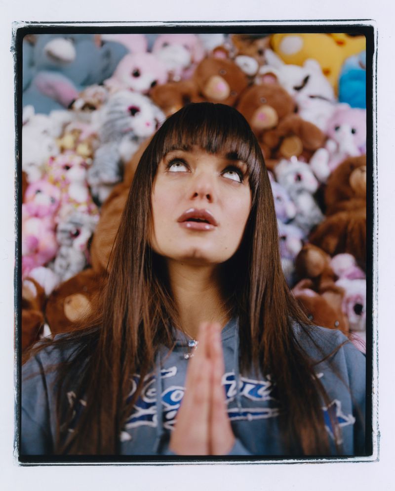 Young woman with bangs, wearing a hoodie, looking upward with her hands together in front of a background of assorted plush toys.