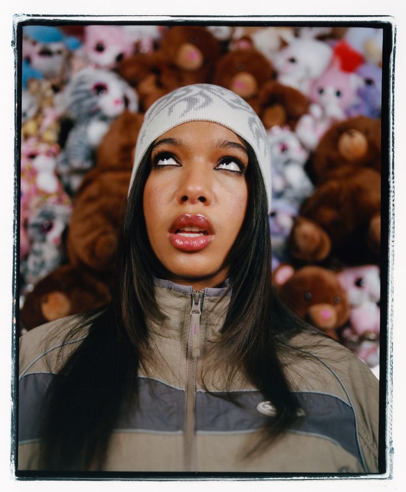 Close-up of an adult female looking upwards, wearing a patterned beanie and beige and grey jacket, against a teddy bear background.