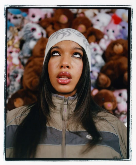 Close-up portrait of a woman wearing a patterned beanie and zip-up jacket against a background of plush toys.
