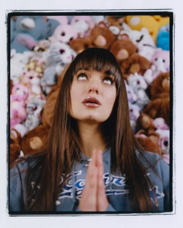 Close-up portrait of a woman with long dark hair and bangs holding hands together in prayer against a wall of stuffed animals.