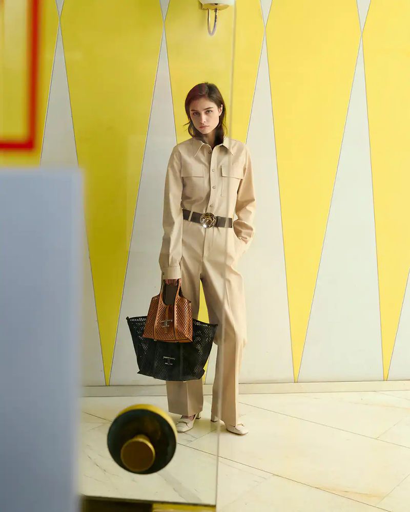 An adult female model poses indoors with a yellow patterned wall behind her, wearing a beige jumpsuit, holding black and tan bags.
