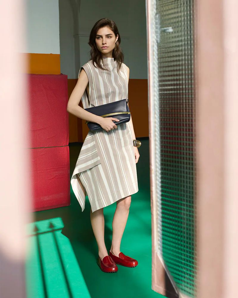A woman models a striped dress with a navy clutch and red loafers. The photo is taken indoors with a blurred foreground and a green floor.
