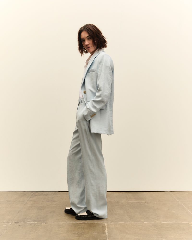 Adult woman in a light blue linen suit poses in a studio. The model is wearing a classic blazer and relaxed wide leg trousers.