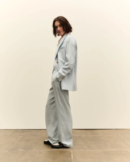 Adult woman in a light blue linen suit poses in a studio. The model is wearing a classic blazer and relaxed wide leg trousers.