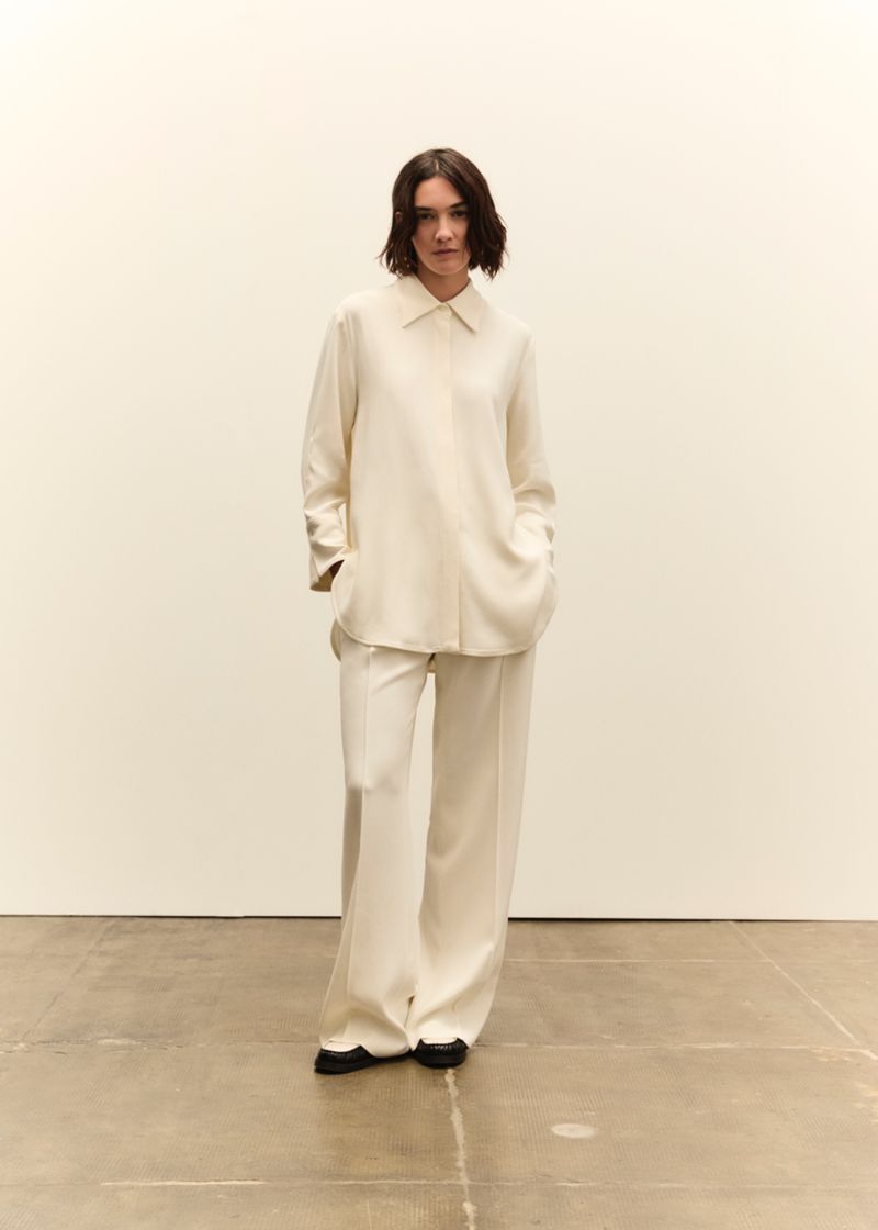 A woman in a cream-colored blouse and wide-leg pants stands in a bright studio. The image has a minimalist aesthetic with neutral tones.