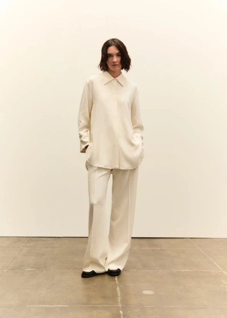 A woman in a cream-colored blouse and wide-leg pants stands in a bright studio. The image has a minimalist aesthetic with neutral tones.