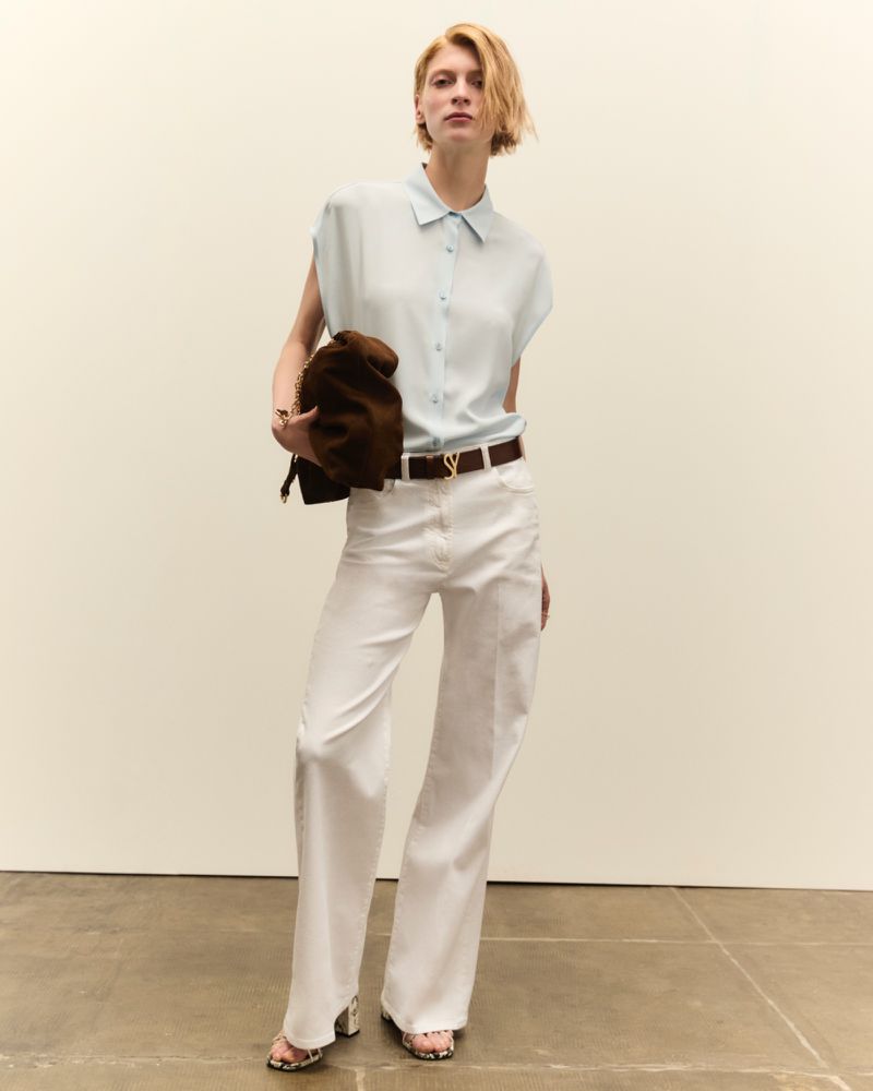 Fashion shot of a young woman in white pants and light blue shirt, holding a bag, posing in a neutral indoor setting with minimal props.
