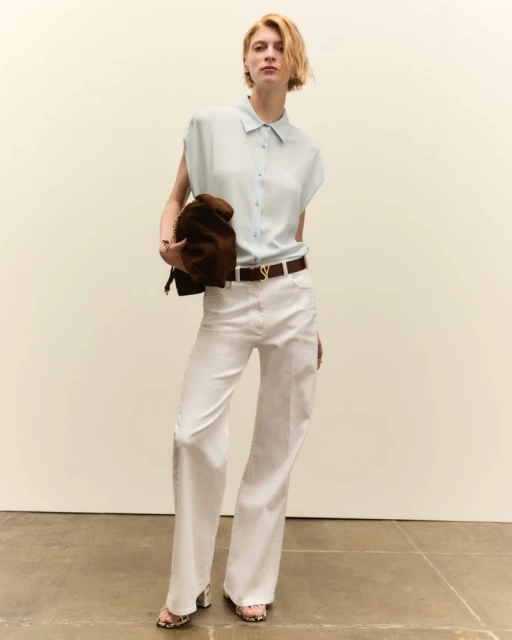 Fashion shot of a young woman in white pants and light blue shirt, holding a bag, posing in a neutral indoor setting with minimal props.