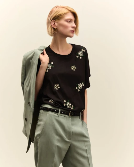 Adult female wearing a black t-shirt with a light green blazer and pants against a neutral backdrop in a studio setting.