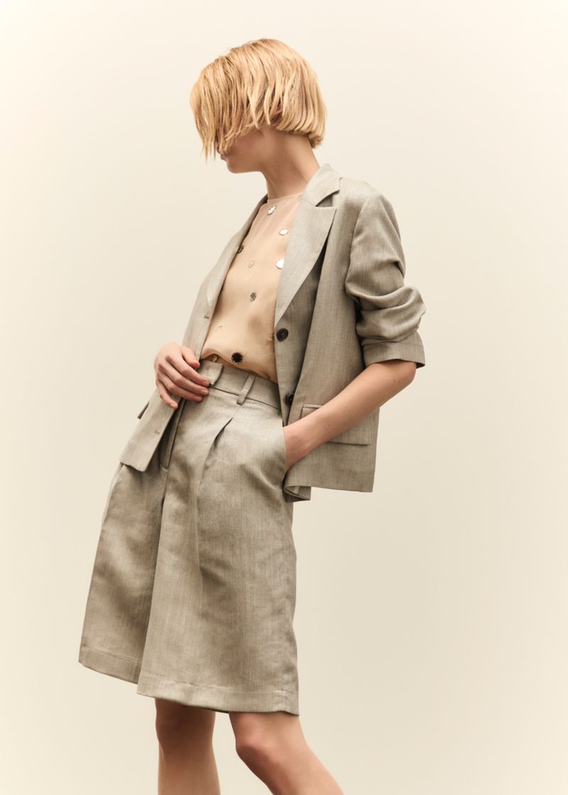Adult female poses in a neutral, indoor setting showcasing a sand-colored linen blazer and matching shorts; fashion editorial style.