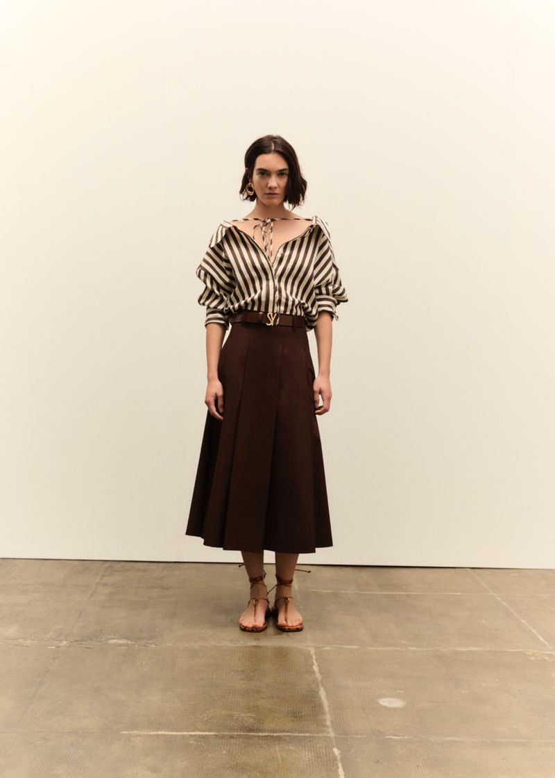Full shot of a woman in a brown and white striped blouse, brown skirt, and sandals against an off-white background in a fashion style.