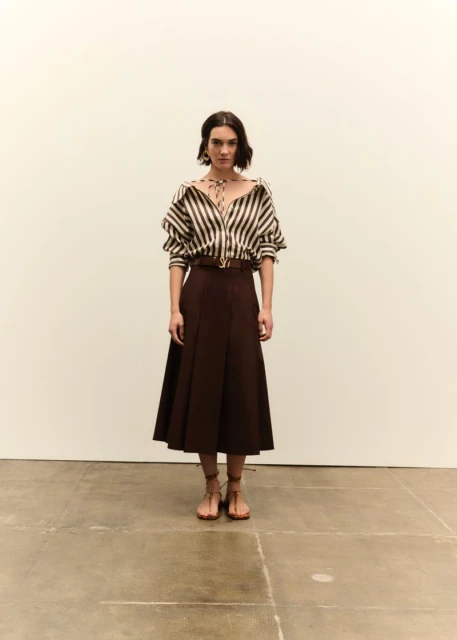 Full shot of a woman in a brown and white striped blouse, brown skirt, and sandals against an off-white background in a fashion style.