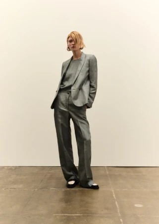 Model wearing a grey trouser suit standing against a plain light wall on a tiled floor