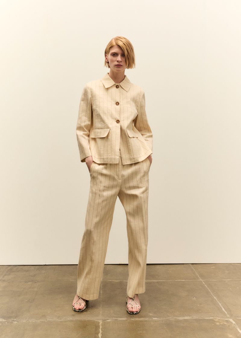 Fashion shot of a redhead woman in a tan striped suit with a blazer and pants, standing in front of a neutral wall indoors.