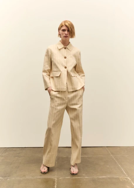 Fashion shot of a redhead woman in a tan striped suit with a blazer and pants, standing in front of a neutral wall indoors.