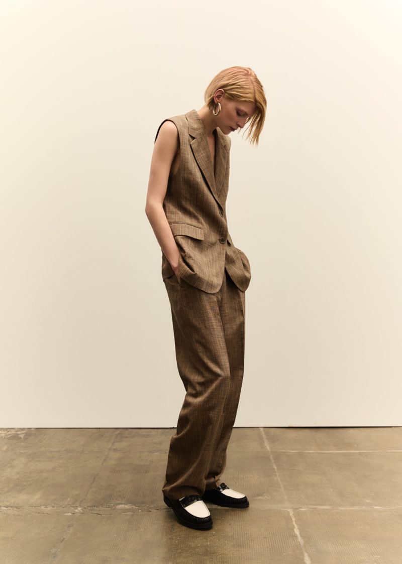 A blond woman stands in a photo studio. She wears a brown vest suit with wide-leg pants and black-and-white loafers, looking down.