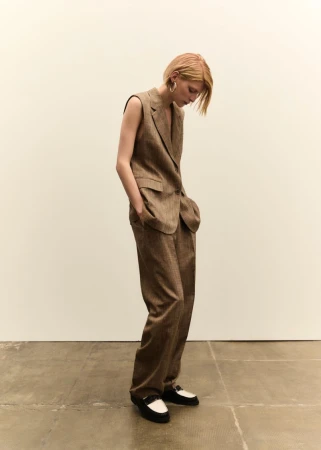 A blond woman stands in a photo studio. She wears a brown vest suit with wide-leg pants and black-and-white loafers, looking down.