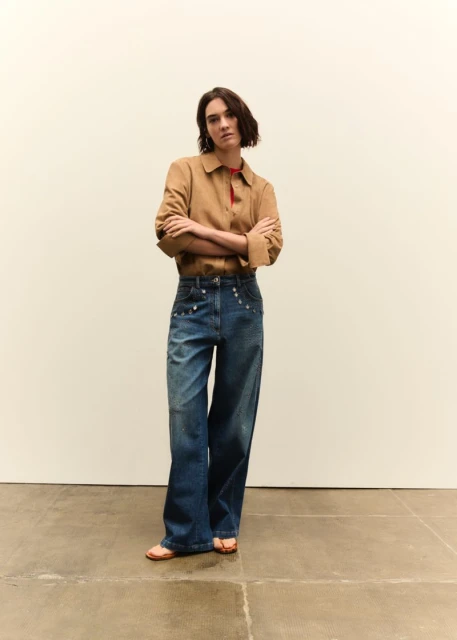 Adult woman poses indoors wearing a tan collared jacket, blue jeans with silver embellishments, and sandals against a cream wall.