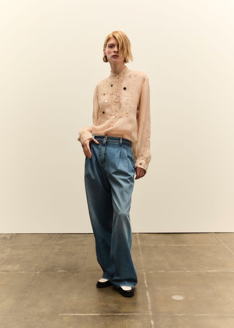 Model in a pale embellished blouse and wide-leg denim trousers standing against a plain white studio background.