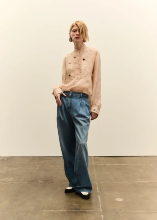 Model in a pale embellished blouse and wide-leg denim trousers standing against a plain white studio background.
