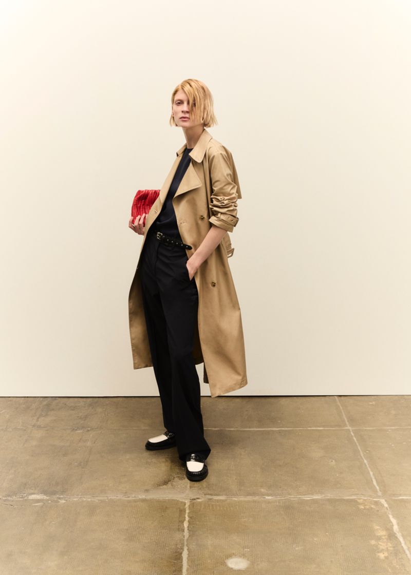 A blond woman in a tan trench coat, black pants, and black and white shoes holds a red clutch against a neutral background.