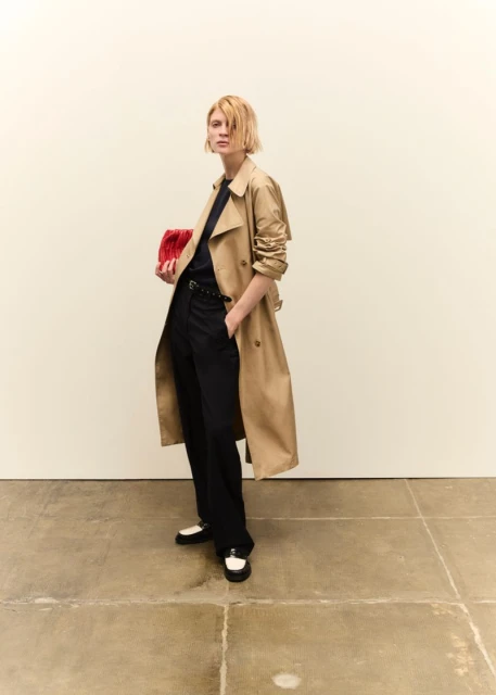 A blond woman in a tan trench coat, black pants, and black and white shoes holds a red clutch against a neutral background.