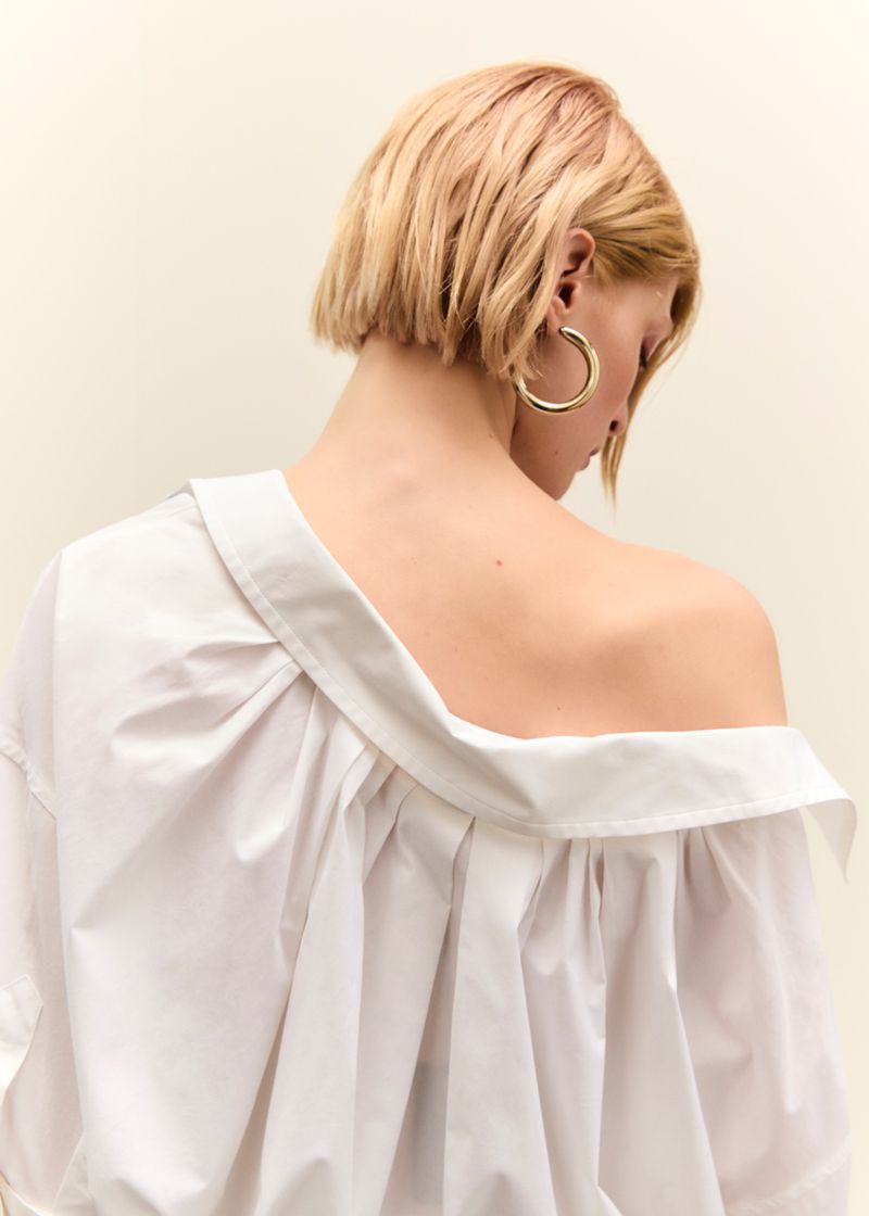 Blonde woman with bare back and exposed shoulder facing away, wearing a white shirt and hoop earring against a cream background.