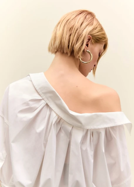 Blonde woman with bare back and exposed shoulder facing away, wearing a white shirt and hoop earring against a cream background.