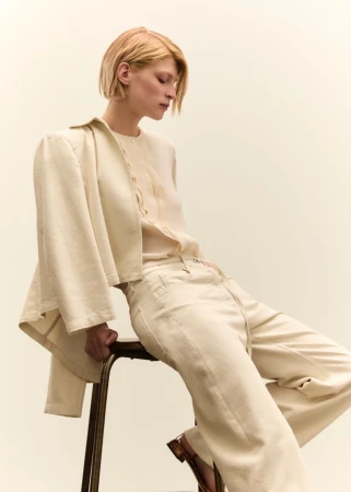 Model in neutral cream clothing sitting on a tall stool against a solid pale background for Seventy Venezia SS25.