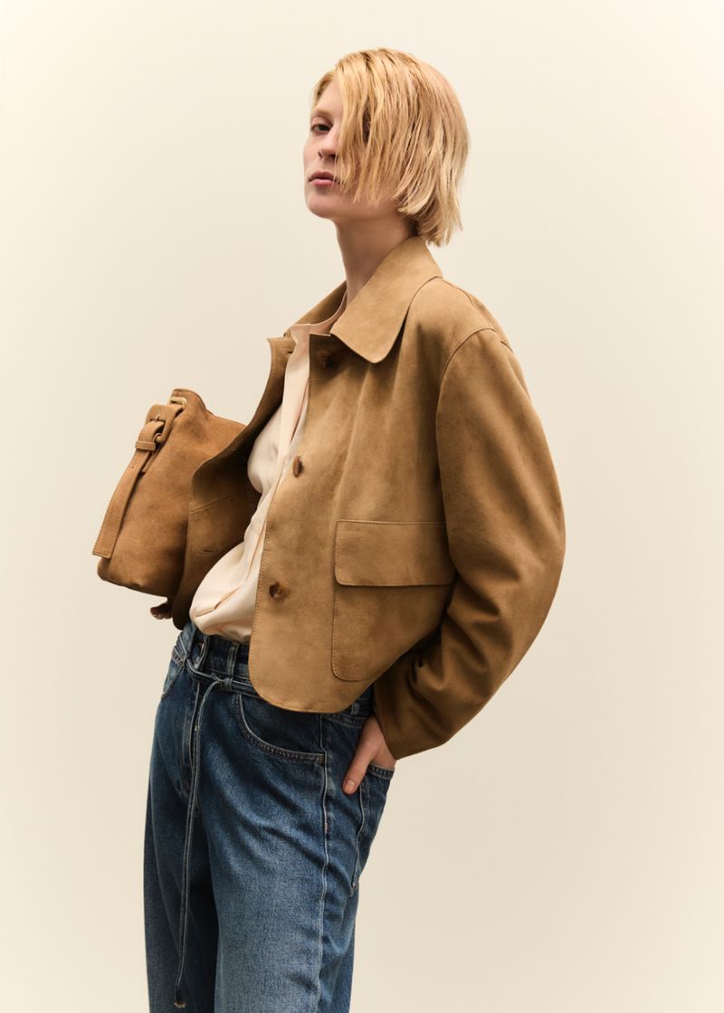 A blonde woman poses indoors wearing a brown jacket, light shirt, and blue jeans, with a brown leather bag over her shoulder.