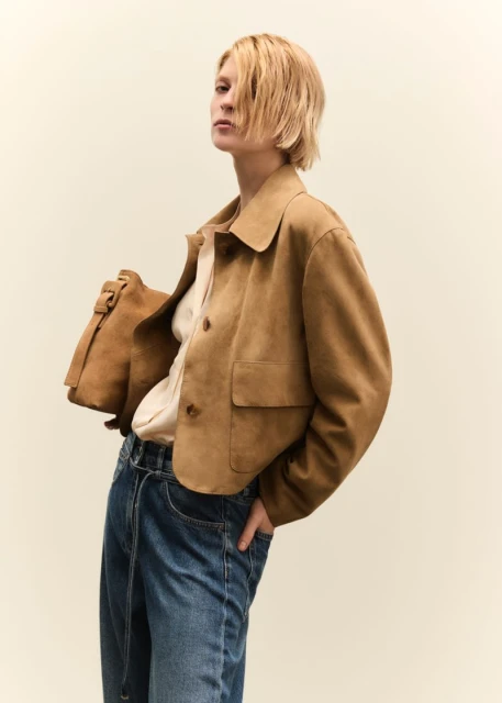 A blonde woman poses indoors wearing a brown jacket, light shirt, and blue jeans, with a brown leather bag over her shoulder.