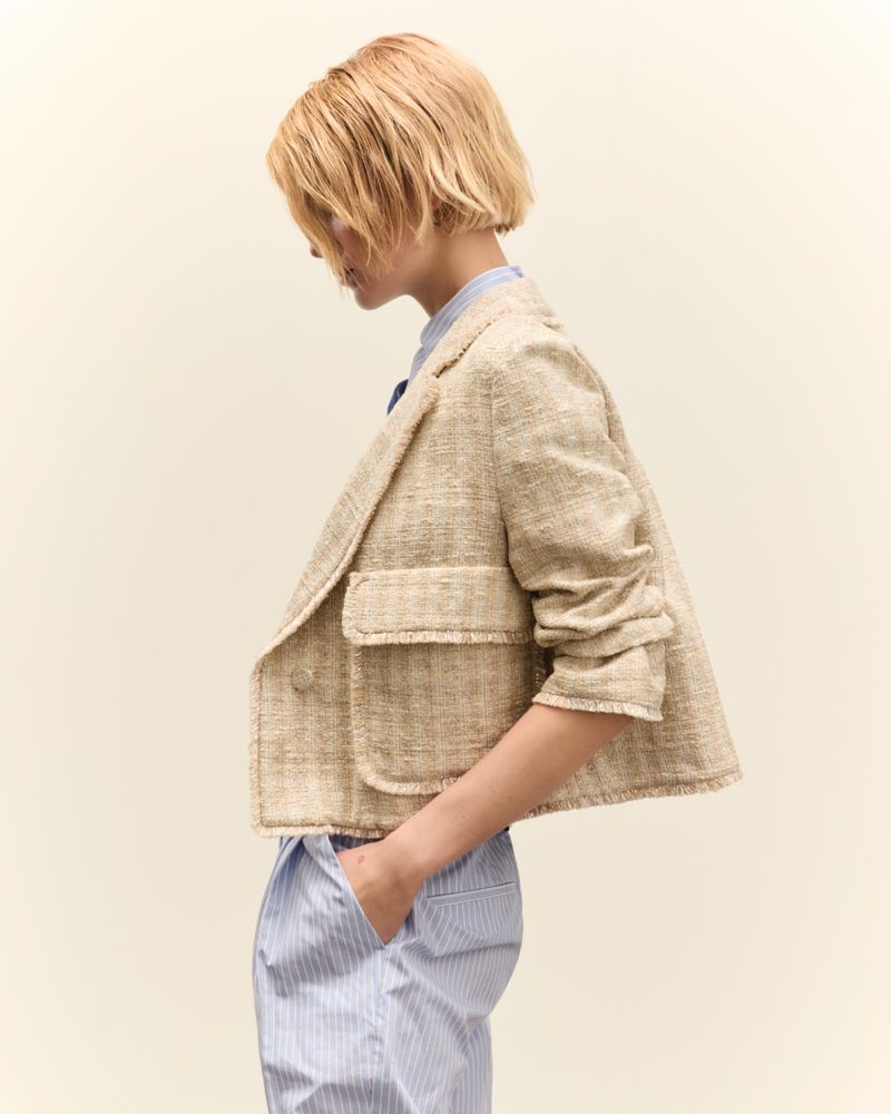Profile shot of a woman in a cropped beige jacket and striped pants against an off-white background, gazing downward in a neutral expression.