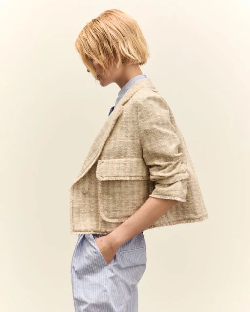 Profile shot of a woman in a cropped beige jacket and striped pants against an off-white background, gazing downward in a neutral expression.
