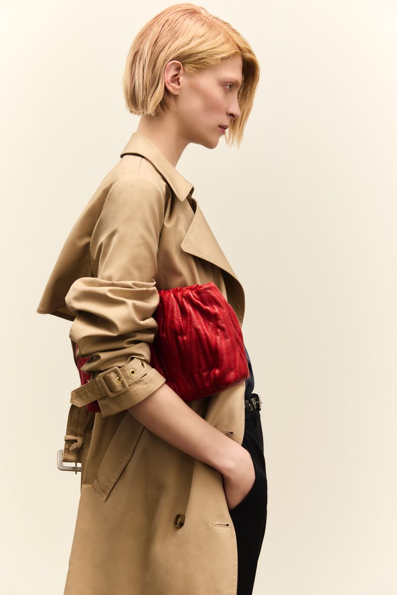 Model with blonde hair wearing a beige trench coat holding a red clutch against a neutral background