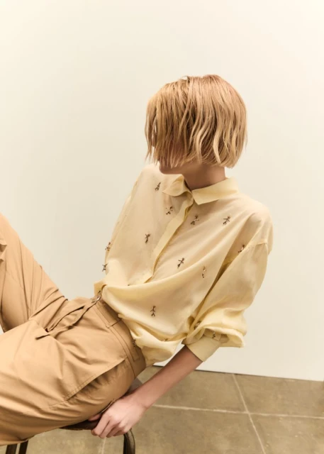 Blonde woman in beige blouse and pants poses in a studio. She's sitting on a stool, looking back over her shoulder in a fashion shot.