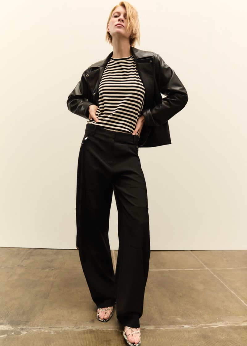Model wearing a black leather jacket, striped top, and black trousers against a neutral background