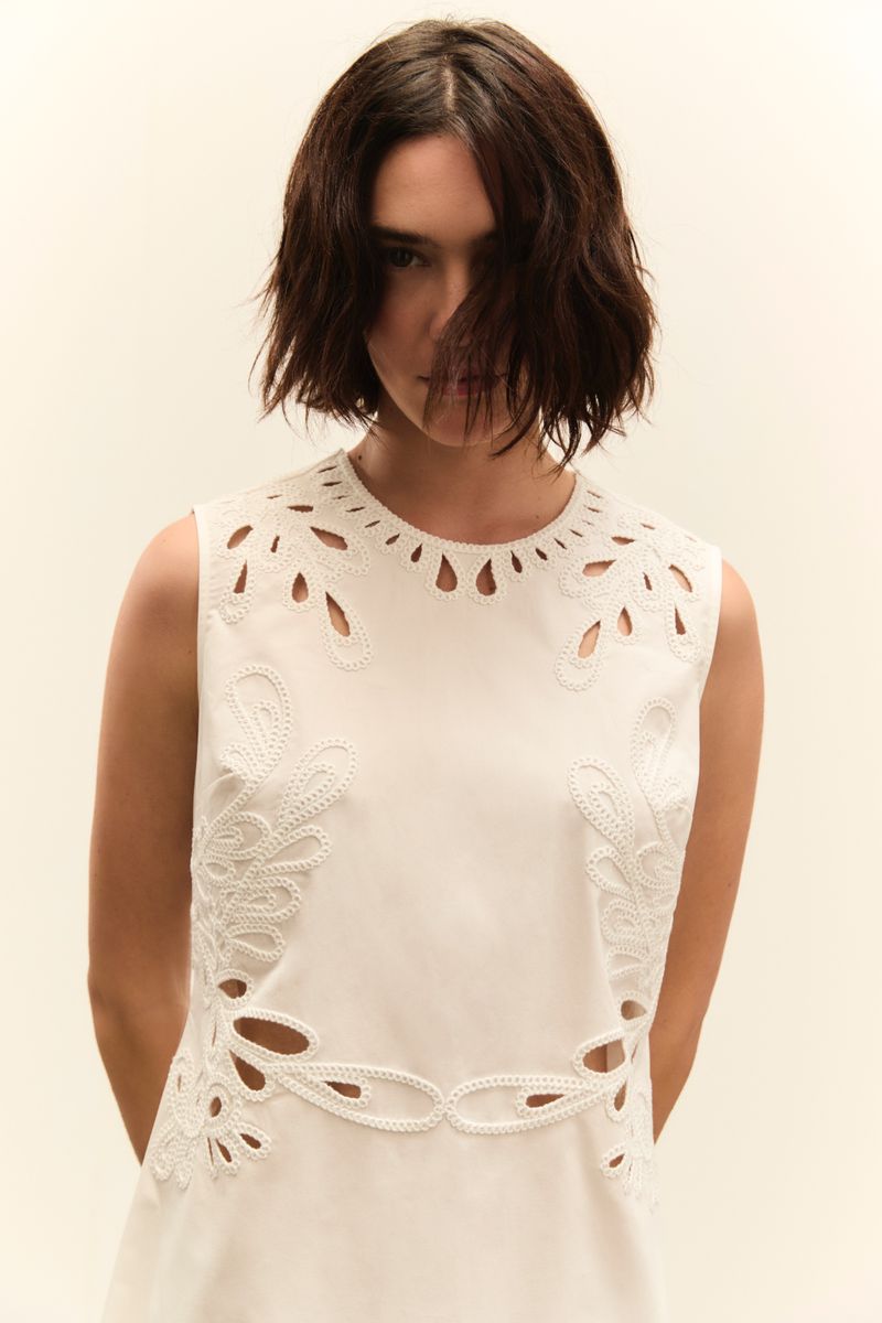 Brunette woman in a sleeveless, knee-length white dress with lace cut-out details posing in front of a soft cream background.