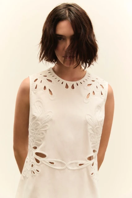 Brunette woman in a sleeveless, knee-length white dress with lace cut-out details posing in front of a soft cream background.