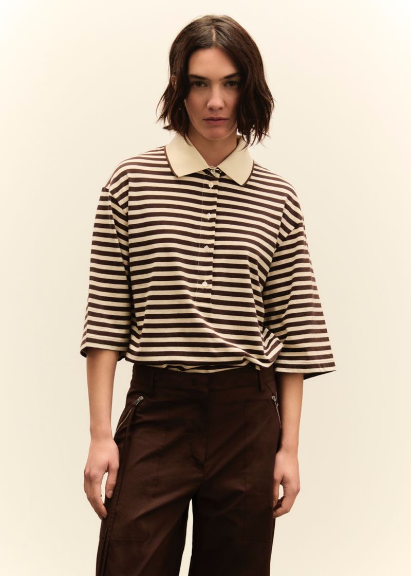 A woman with short dark hair is centered in a medium shot, wearing a tan and brown striped shirt and dark brown pants, against a neutral backdrop.