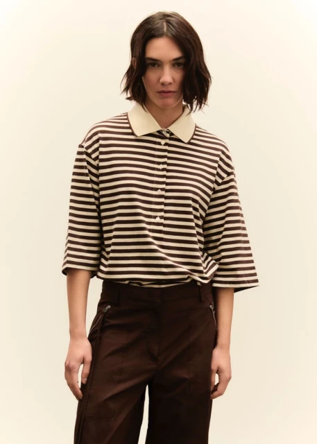 A woman with short dark hair is centered in a medium shot, wearing a tan and brown striped shirt and dark brown pants, against a neutral backdrop.