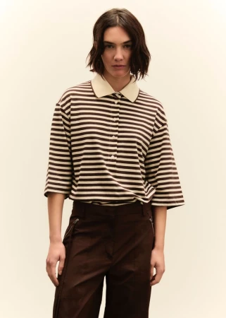 A woman with short dark hair is centered in a medium shot, wearing a tan and brown striped shirt and dark brown pants, against a neutral backdrop.