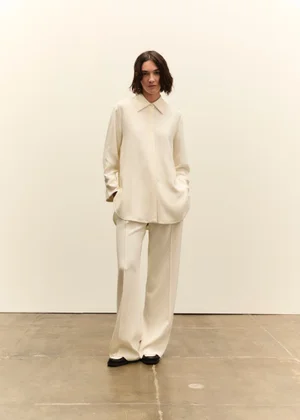 Model wearing a cream button-down shirt and matching trousers standing against a pale neutral background