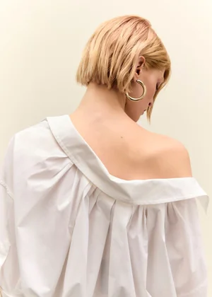 Model with blonde bob hair wearing an off-the-shoulder white blouse and gold hoop earrings against a plain background