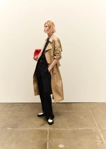 Model in camel trench coat and black trousers holding a red clutch standing against a plain wall.
