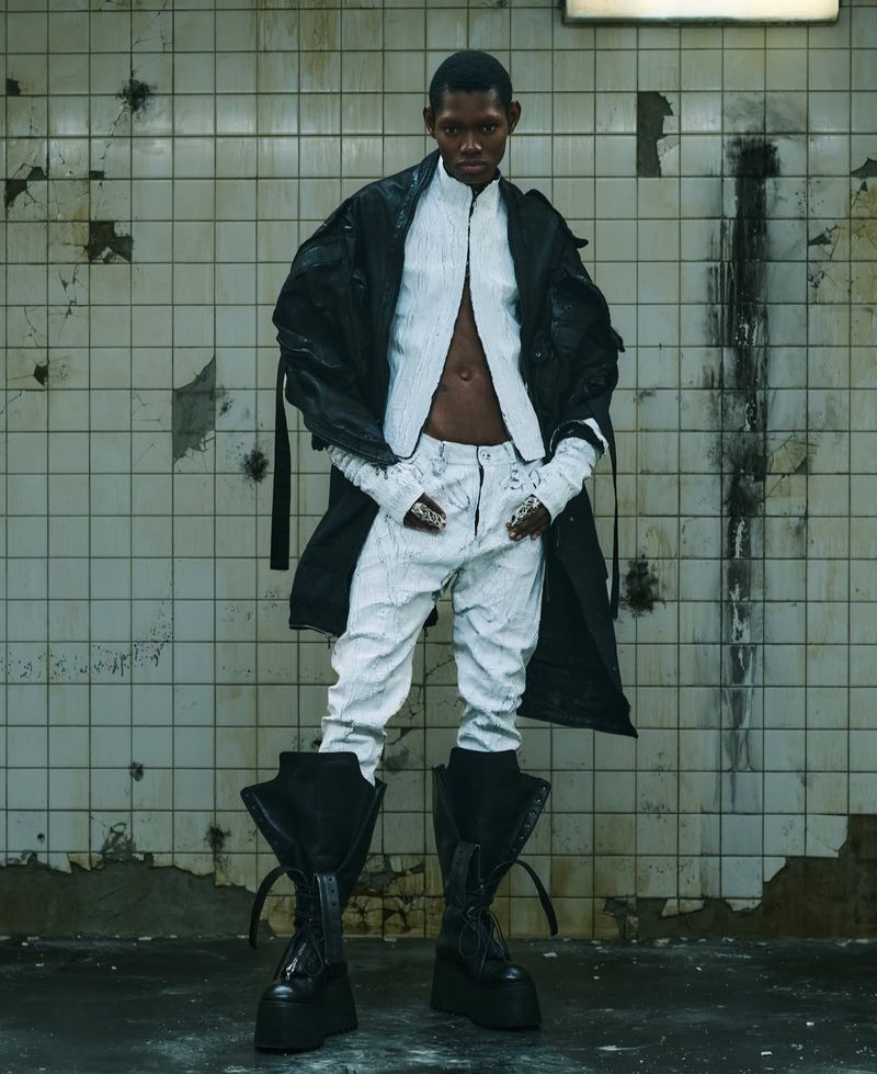 A slender Black adult stands in a gritty urban setting wearing a modern fashion ensemble with large boots, a long black coat, and white garments.