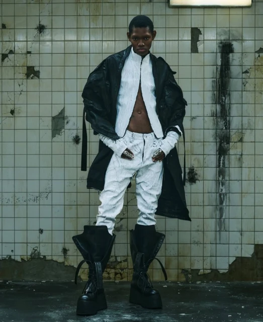 A slender Black adult stands in a gritty urban setting wearing a modern fashion ensemble with large boots, a long black coat, and white garments.
