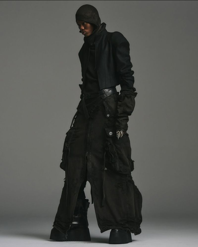 A model poses in a dark, avant-garde ensemble featuring a cropped jacket, layered pants, and platform shoes against a solid grey backdrop.