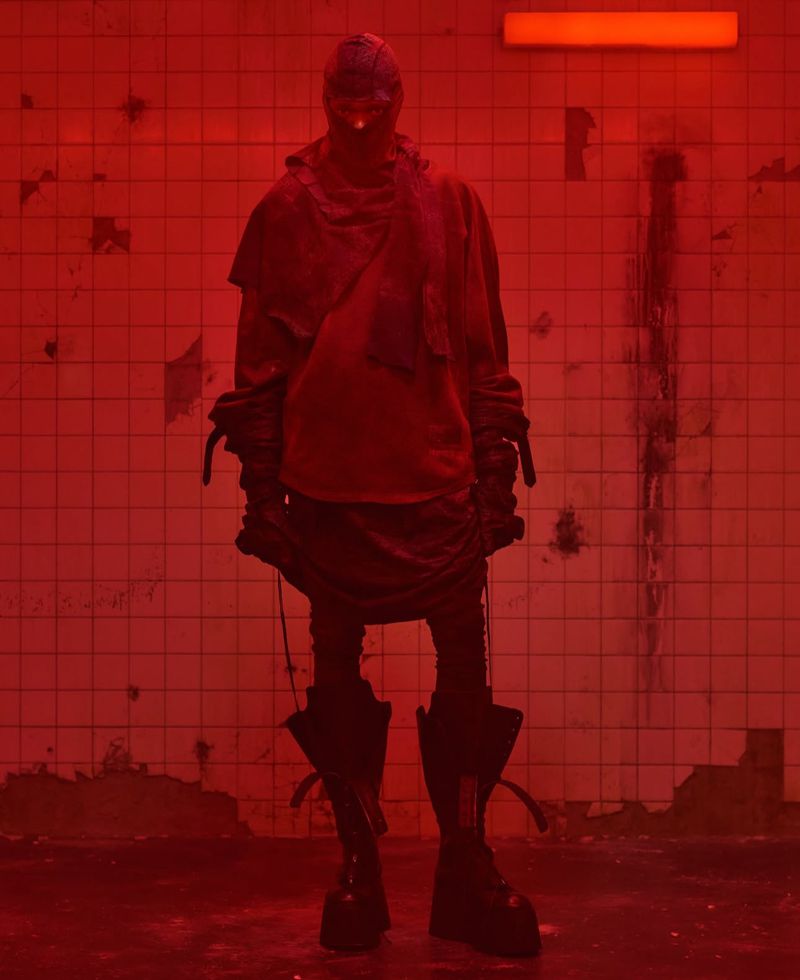 Figure in a red-lit space wearing a hooded garment with straps and boots. The scene is framed against a gridded tile wall.